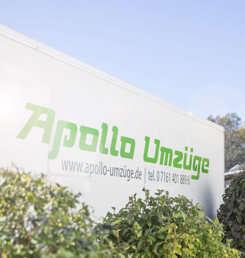Seitenansicht eines weißen Umzugswagens mit grünem Schriftzug Apollo Umzüge, teilweise von Büschen verdeckt.