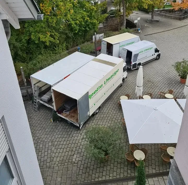 Zwei Lieferwagen auf gepflastertem Hof neben Tischen mit Stühlen und großen Sonnenschirmen.