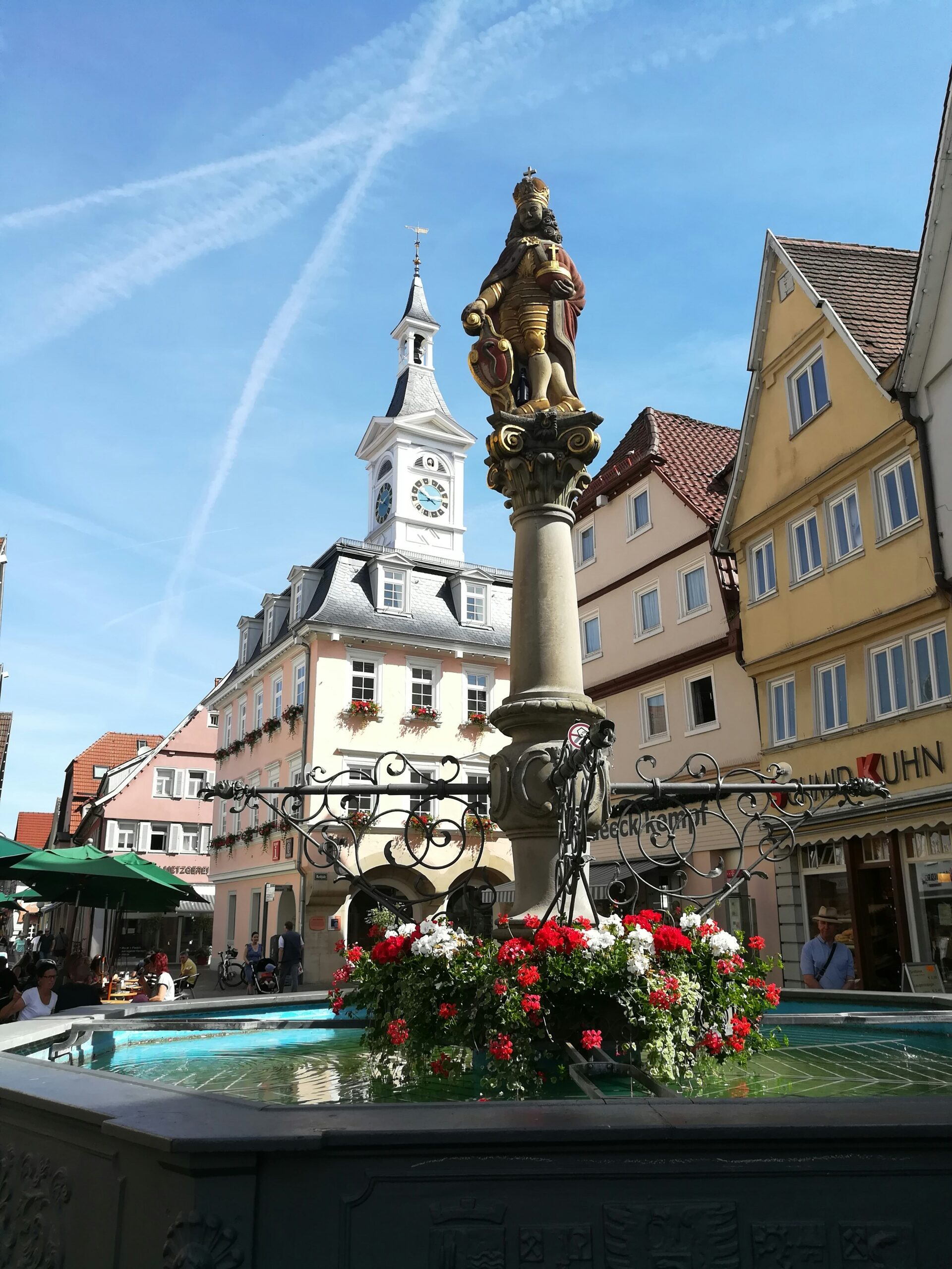 Brunnen mit Statue in einem historischen Stadtplatz mit Fachwerkhäusern und Uhrturm bei klarem Himmel.