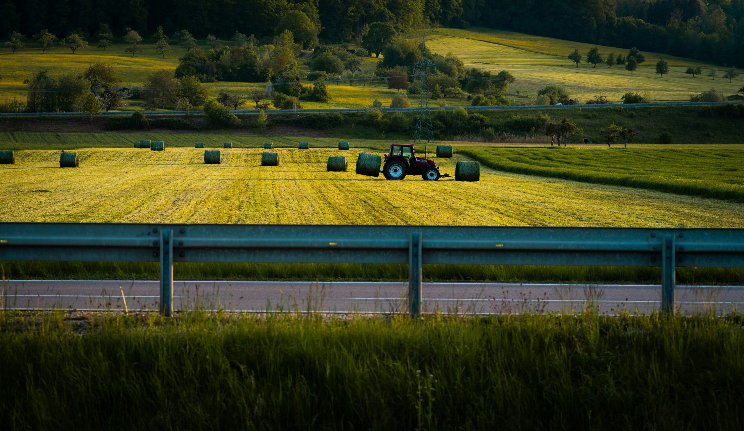 Traktor wickelt Heuballen auf einem Feld bei Sonnenuntergang, im Vordergrund eine Straße mit Leitplanke.