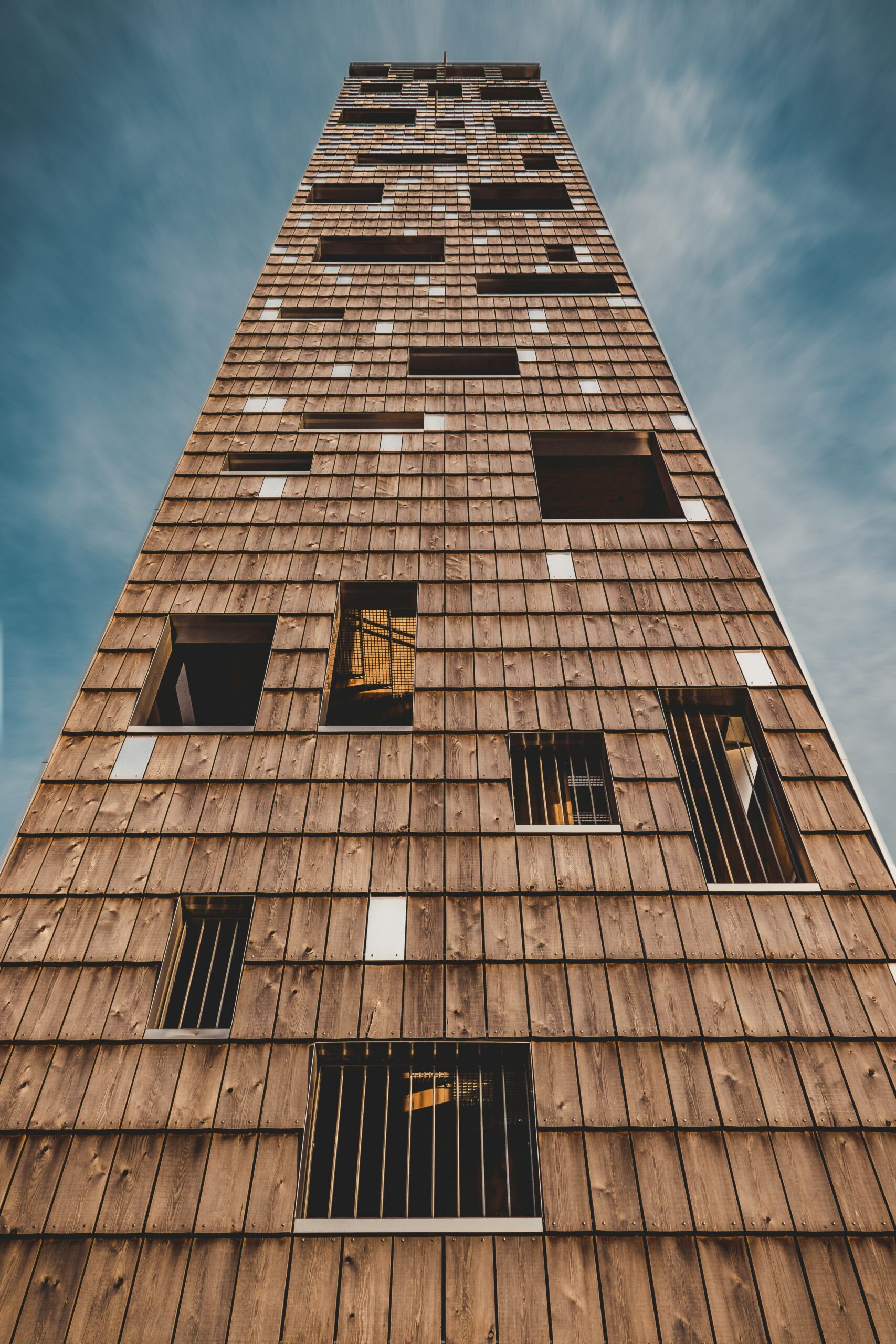 Holzturm mit unregelmäßig verteilten Fenstern vor bewölktem Himmel.