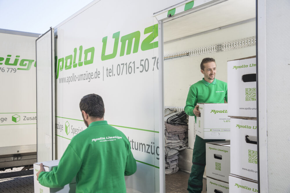 Zwei Männer in grünen Pullovern laden Umzugskartons in einen weißen Umzugswagen.