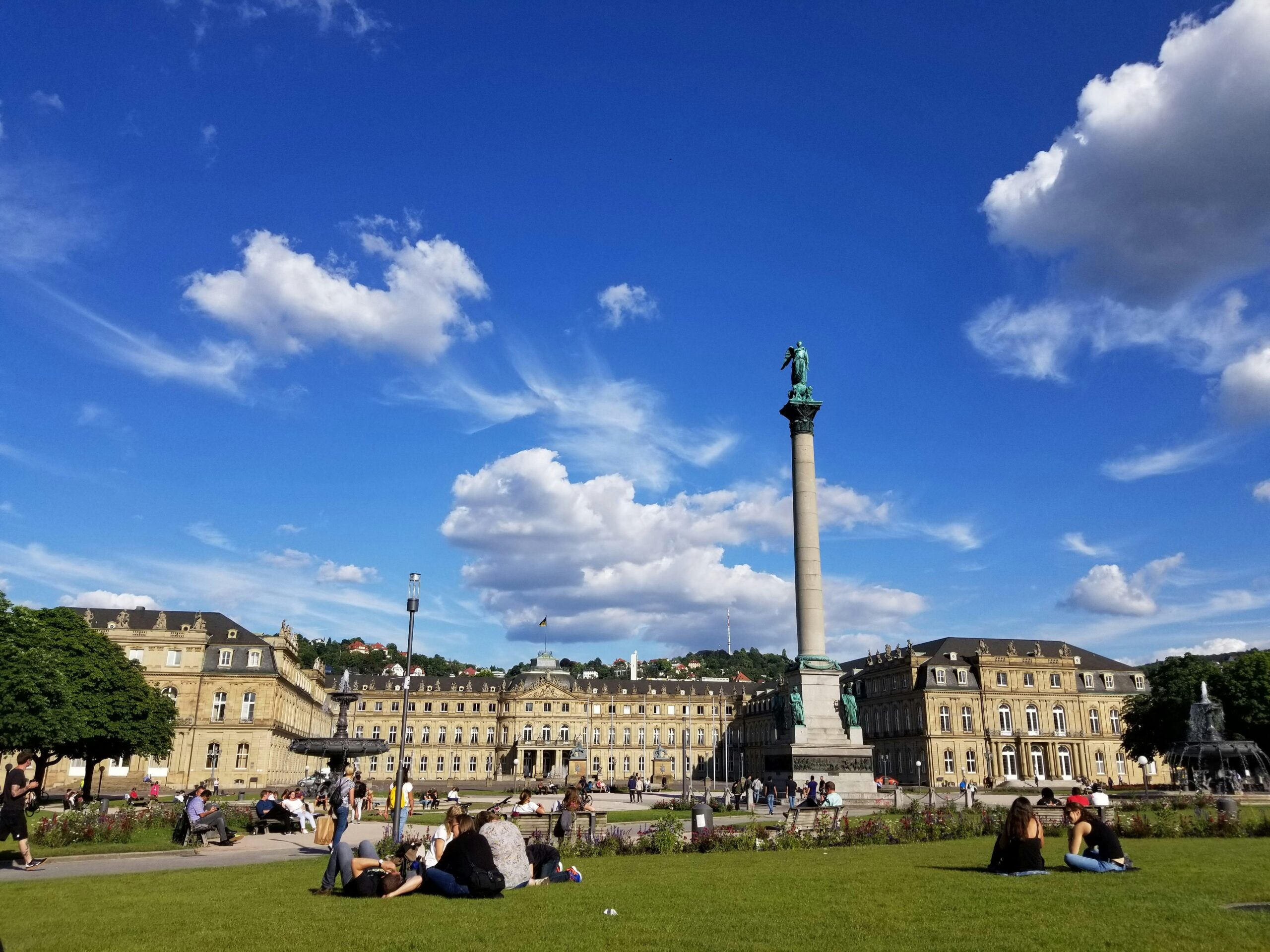 Menschen sitzen auf einer Wiese vor einer historischen Säule mit Statue und Gebäuden unter blauem Himmel mit Wolken.