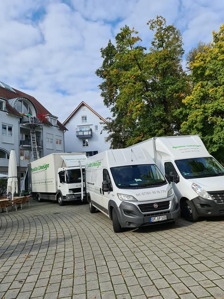 Drei weiße Lieferwagen vor einem Gebäude mit Bäumen und bewölktem Himmel.